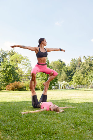 Two young sportswomen doing acrobatic yoga or yoga partner together outdoors in fitness park....