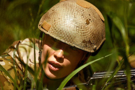 Caucasian Military lady woman in tactical gear posing for photo at summer season. Wearing green camo uniform and assault rifle, in military gear and headset, lady is looking at sideの写真素材