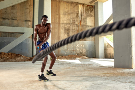 Rope workout. Sport man doing battle ropes exercise outdoor. Black male athlete exercising, doing...