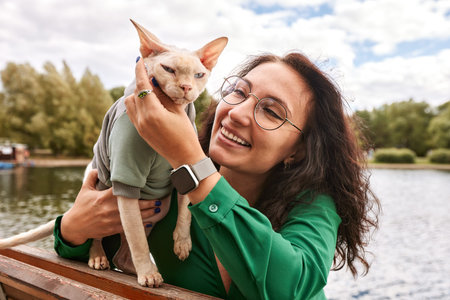 Cute girl petting a furry cat outdoor. Sitting on wooden bench with green trees and river...