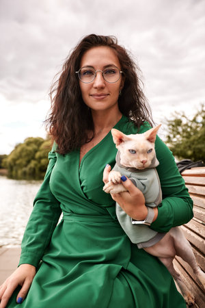Cute girl petting a furry cat outdoor. Sitting on wooden bench with green trees and river background. Love to the animals.の写真素材