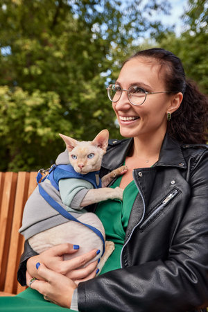 Cute girl petting a furry cat outdoor. Sitting on wooden bench with green trees and river...