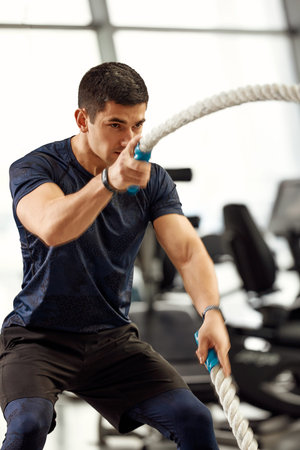 young handsome man in a black t-shirt is engaged with ropes in the gym on a window background.