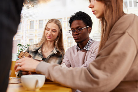 Multiracial happy young people, black and white cheerful comrades laughing, enjoying food, having fun sitting together at a table in a restaurant, different friends sharing lunch at a meetingの写真素材