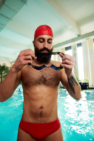 A swimmer in a cap and goggles puts on swimming goggles smiling before swimming training in the pool. Training, training or exercises for athletes in swimming for fitness or health purposesの写真素材