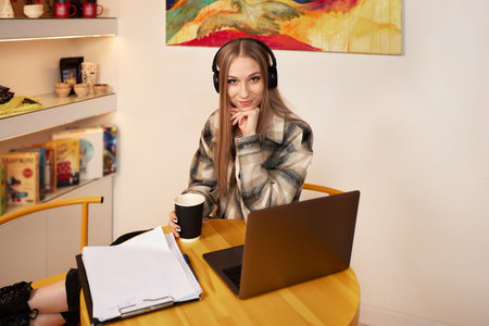 Young smiling female student using laptop for e-learning in headphones, watching online seminar in...