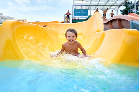 Cheerful blue-eyed kid on a water slide in the water park, a little boy merrily slides into the water along the slide in the amusement park, rest with children in the water parkの写真素材