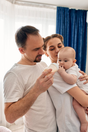 Baby on parents arms drinks baby food from bottle. Mom and dad keep care of their adorable infant...