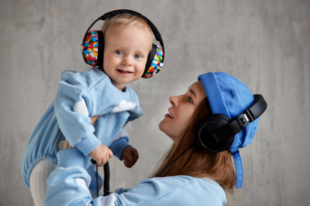 A happy young mother hugs her smiling baby and lifts him up in her arms. Baby and mom in blue clothes and DJ headphones on their heads.の写真素材