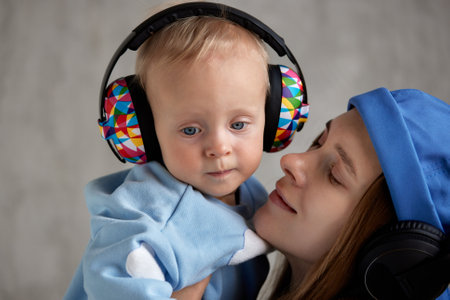 A happy young mother hugs her precious baby and cuddles her. Baby and mom in blue clothes and DJ headphones on their heads.の写真素材