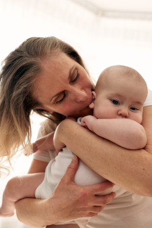 Mom hugs and kisses her little child in a bright childs room. Mom carefully holds the baby in her arms. Happy childhoodの写真素材