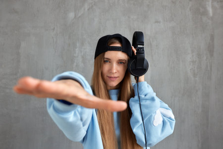 A young pretty long-haired DJ girl in a blue sweater and a black baseball cap holds headphones in her hand and imitates the movements of a hip-hop musician. Music, fun and beauty.の写真素材
