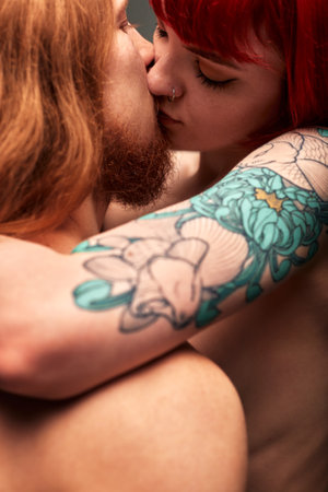 A young attractive woman with bright ruby hair and a tattoo hugs and kisses a man with long hair. Half-naked lovers in a passionate kiss. Close-up studio shot, dark background.の写真素材