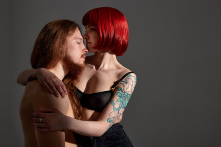 A young attractive woman with bright ruby hair and a tattoo in black lingerie embraces a man with long hair and a beard. Half-naked lovers in each others arms. Close-up studio shot, dark background.の写真素材