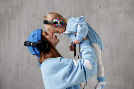 A happy young mother hugs her smiling baby and lifts him up in her arms. Baby and mom wear blue clothes and have funny DJ headphones on their heads. Studio shot, grey background.の写真素材