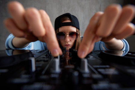 A young pretty long-haired DJ girl in a blue sweater and black baseball cap poses with a black DJ mixing console and mixes music tracks. Close-up studio shot, gray background.の写真素材