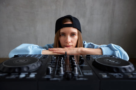 A young pretty long-haired DJ girl in a blue sweater and black baseball cap poses with a black DJ mixing console and mixes music tracks. Close-up studio shot, gray background.の写真素材