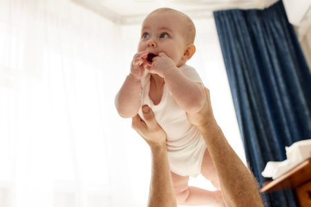 A cute joyful smiling baby on the outstretched arms of his father, who lies on the floor in a bright room. Happy fatherhood and childhood.の写真素材