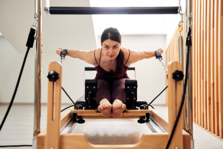 A young girl is doing Pilates on a reformer bed in a bright studio. A slender brunette does exercises to strengthen the muscles of her arms and shoulder girdle. Healthy lifestyle concept.の写真素材