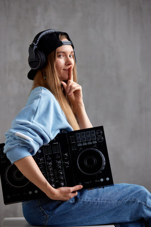 A young pretty long-haired DJ girl in a blue sweater, jeans and a black baseball cap poses while sitting with a black mixing console in her hand. Studio shot, gray background.の写真素材