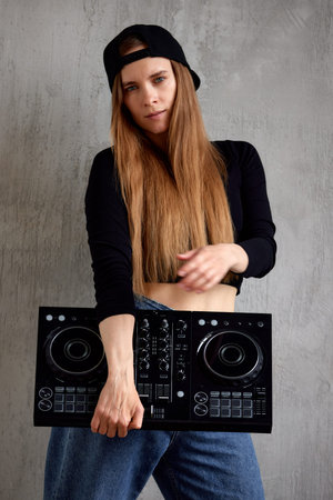 A young pretty long-haired DJ girl in a black top, blue loose jeans and a black baseball cap stands...