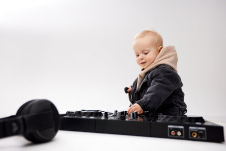 A cute kid in a beige hoodie and a funny black motorcycle jacket sits on the floor with dj headphones and a dj mixing console. Music and fun. Isolate on a white background.の写真素材