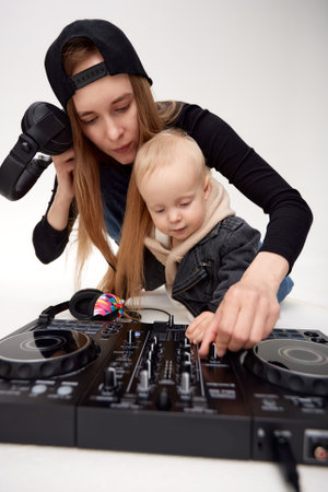 A young mother in a black baseball cap and her toddler in a funny black motorcycle jacket with DJ headphones are using a DJ mixing console. Music and fun. Isolate on a white background.の写真素材