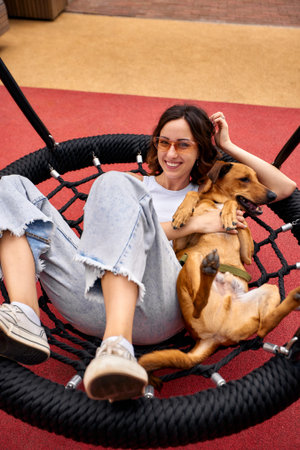 Charming young smiling girl is resting on a swing in the city yard with a golden dog on a sunny day. The girl hugs her pet. Love and affection between owner and pet. Adopting a pet from a shelter.の写真素材