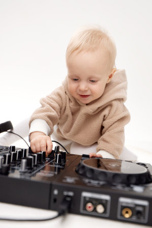 A cute smiling blonde baby in a beige hoodie sits on the floor and plays with dj headphones and a dj mixing board. Music and fun. Isolated on a white background.の写真素材