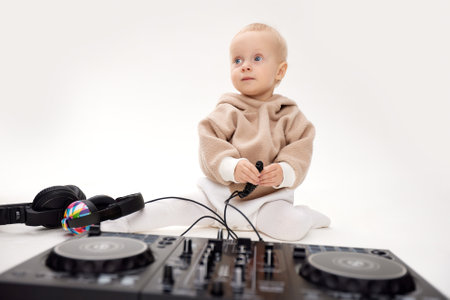 A cute smiling blonde baby in a beige hoodie sits on the floor and plays with dj headphones and a dj mixing board. Music and fun. Isolated on a white background.の写真素材