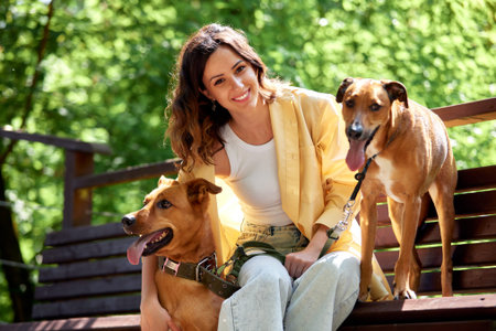 A young charming smiling girl is resting on a bench while walking in the park with two golden dogs....