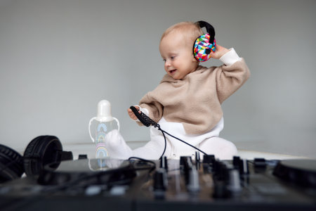 A cute smiling blonde baby in a beige hoodie sits on the floor and plays with dj headphones and a dj mixing board. Music and fun. Isolated on a gray background.の写真素材