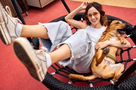A young charming smiling girl is resting on a swing in the city yard with a golden dog. The girl hugs her pet and plays with it. Love and affection between owner and pet.の写真素材