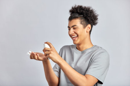 Young handsome smiling guy with black curly hair squeezing skincare cream from the dispenser to his hand. Biracial man in studio with healthy skin procedure. Wellness, cosmetics and skincare for men.の写真素材