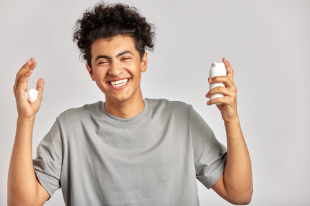 Young handsome smiling guy with black curly hair squeezing skincare cream from the dispenser to his hand. Biracial man in studio with healthy skin procedure. Wellness, cosmetics and skincare for men.の写真素材