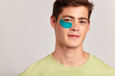 A millennial guy with brown hair practicing skincare with under eye hydrogel patches in a studio. Young caucasian man smiling, absorbing the ingredients of the cosmetic under eye mask. Mens skincare.の写真素材