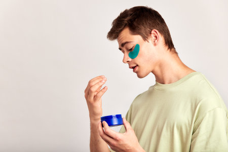 A millennial guy with brown hair practicing skincare with under eye hydrogel patches in a studio. Young caucasian absorbing the ingredients of the cosmetic under eye mask. Mens self-care.の写真素材