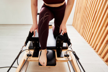 A young girl is doing Pilates on a reformer bed in a bright studio. A slender brunette in a burgundy bodysuit does exercises to strengthen the arms and shoulder girdle. Healthy lifestyle concept.の写真素材