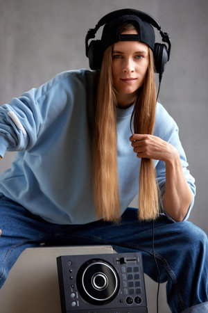 A young pretty long-haired DJ girl in a blue sweater, jeans, a black baseball cap and black headphones sits with a DJ mixing console. Studio shot, gray background.の写真素材