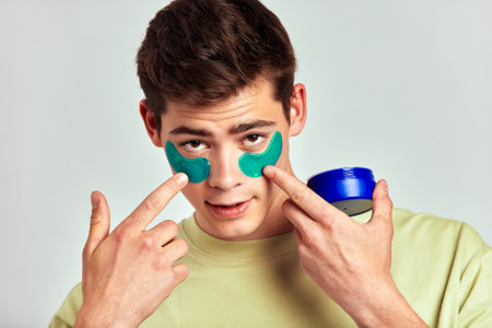 A millennial guy with brown hair practicing skincare with under eye hydrogel patches in a studio. Young smiling caucasian man applying cosmetic patches on his face. Mens self-care.の写真素材