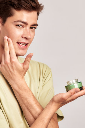 Young guy holding a vial of facial mask cream in his hands and applying it on his face during his skincare routine. Man caring for the skin to maintain his youthful and healthy appearance.の写真素材