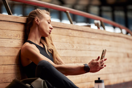 Young athlete girl is having break after a workout in the stadium. A slender athletic smiling blonde in a black tracksuit sitting in a relaxed pose and making selfie. Sports and recreation.の写真素材