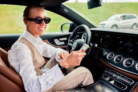 A young attractive business lady dressed in a white shirt and a beige vest is driving her luxury...