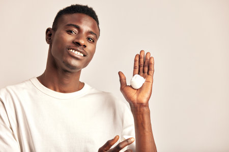 Young handsome smiling African guy with a dash of moisturizing facial cream on his palm in a studio. A black man caring for the skin to maintain his youthful and healthy appearance. Mens self-care.の写真素材