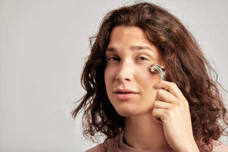 A handsome guy with long brown hair doing self-care routine using a face massage roller in studio. Caucasian man caring for the skin to maintain his youthful and healthy appearance.の写真素材