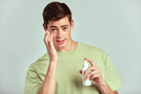 A young handsome guy with brown hair doing self-care routine using a facial moisturizer in a studio. Caucasian man caring for the skin to maintain his youthful and healthy appearance. Mens skincare.の写真素材