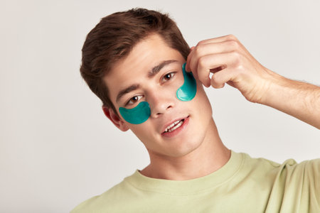 A millennial guy with brown hair practicing skincare with under eye hydrogel patches in a studio. Young smiling caucasian man applying cosmetic patches on his face. Mens self-care.の写真素材