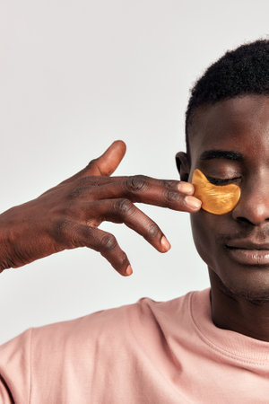 African millennial guy practicing skincare and grooming with under eye hydrogel patches in a studio. Young smiling black man applying cosmetic patches on his face to maintain healthy appearance.の写真素材