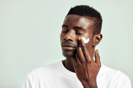 Black guy with youthful skin applying a moisturizing cream on his face to keep his skin soft and smooth. Handsome young African man maintaining his natural beauty with a skincare routine.の写真素材