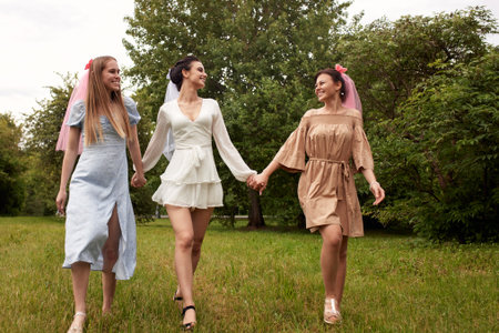A happy charming bride in a white dress with a white veil and two bridesmaids in a blue and beige dress with pink veils walk in a summer park holding hands. Cheerful bachelorette party.の写真素材